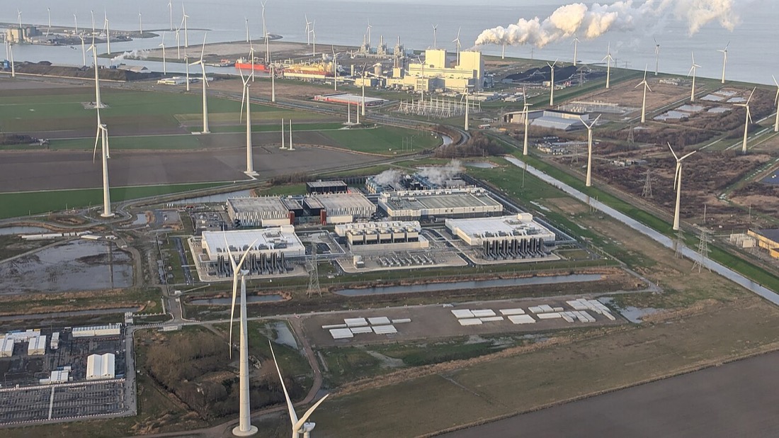 An aerial photograph of a data centre of several long, low buildings in flat Dutch farmland near the sea, with rows of wind turbines around it and a power station with plumes of steam in the background.
