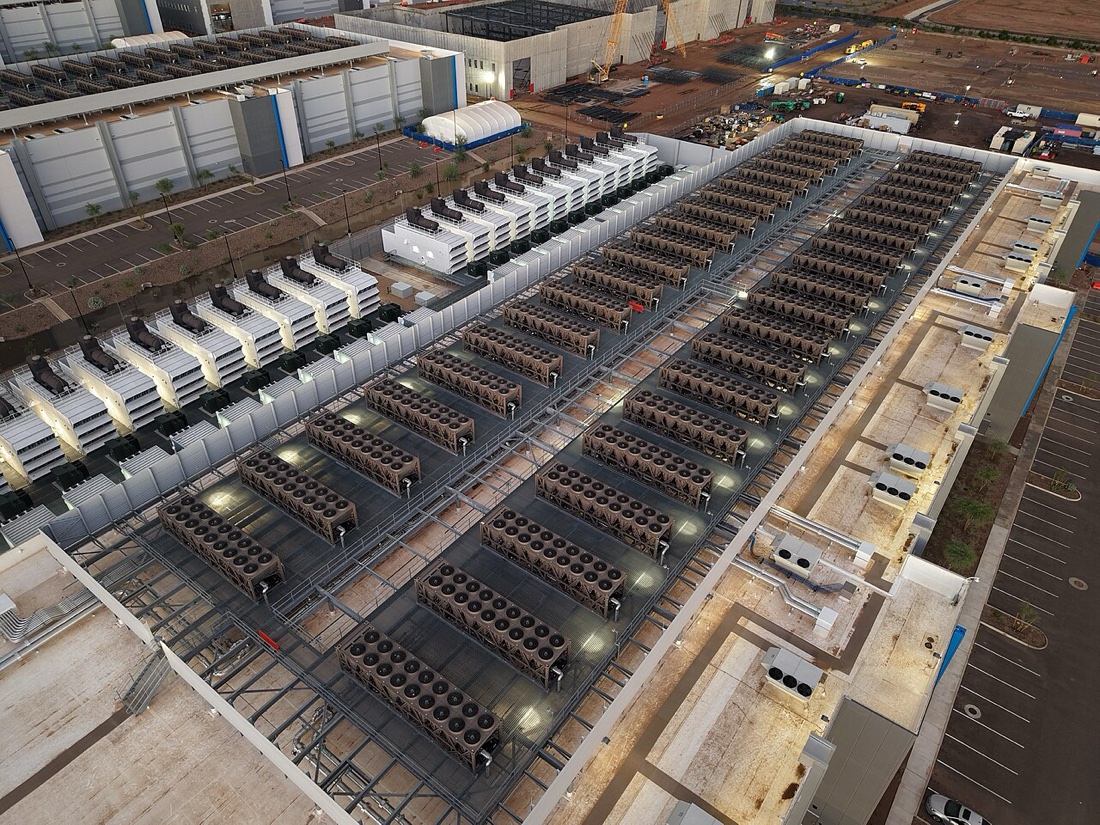 An aerial photograph at dusk of a data centre roof covered in long rows of large fan units, with rows of white generator enclosures to the left, beside a car park and more data-centre buildings under construction.