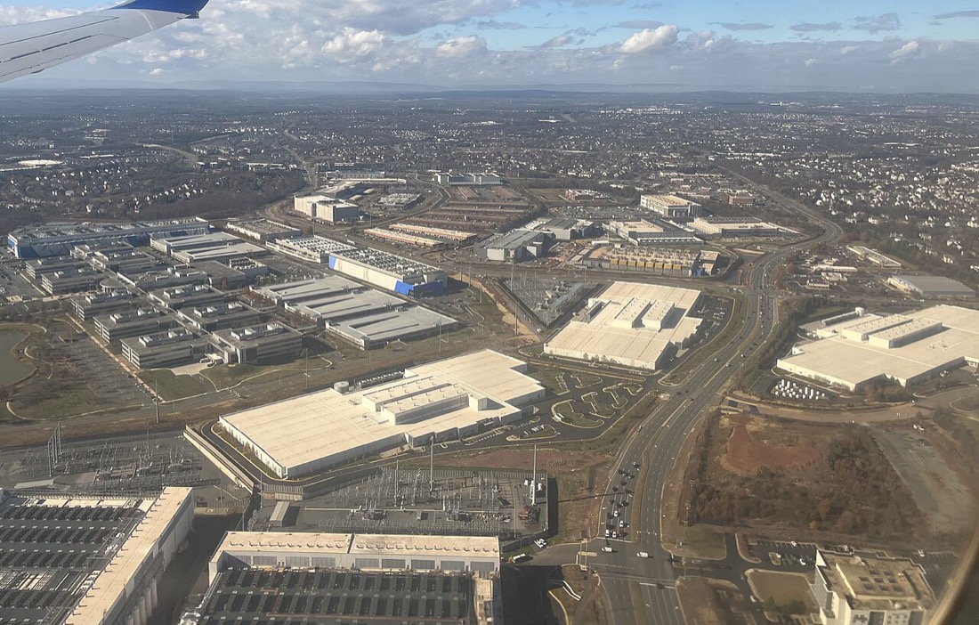An aerial photograph taken from a plane. Large flat-roofed, windowless buildings, several the size of city blocks, stand among roads, car parks and electrical substations, with housing and fields stretching to the horizon.